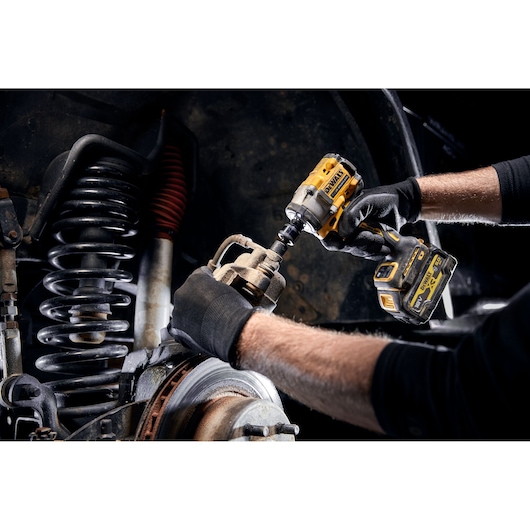 A person wearing black gloves is using a yellow DEWALT cordless impact wrench to work on the suspension and brake system of a vehicle.