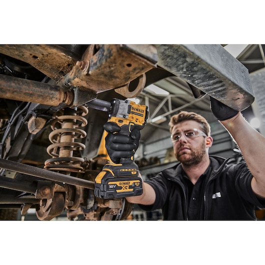 A person wearing black gloves is using a DEWALT DCF923P2G cordless impact wrench to work on the suspension of a vehicle in a workshop. The person's face is blurred for privacy.
