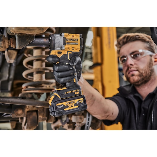 A person wearing black gloves uses a DEWALT 18V XR cordless impact wrench on a vehicle's suspension component. The background shows automotive workshop equipment. The person's face is blurred.