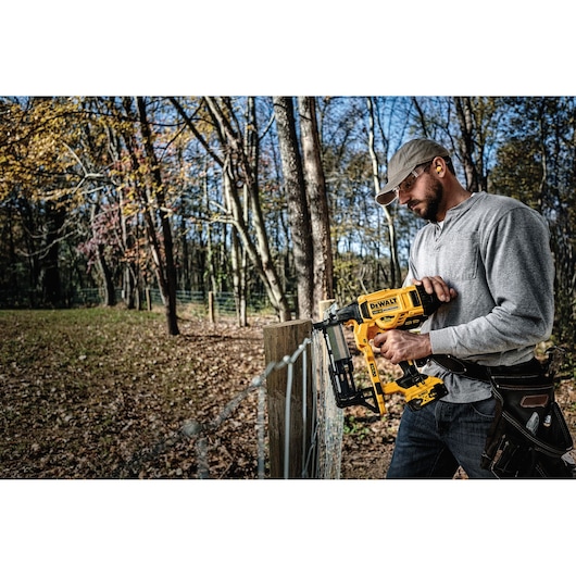 Person using DEWALT cordless fencing nailer to build a wooden fence outdoors in a forested area.