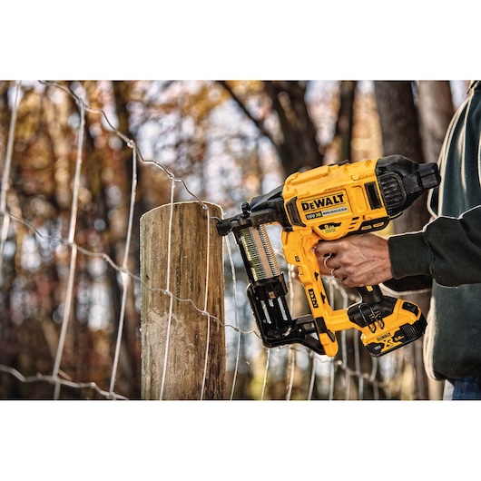 A person uses a yellow DEWALT cordless fencing stapler to attach wire fencing to a wooden post outdoors.
