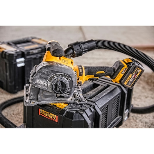 A close-up image of a DEWALT cordless power tool, specifically a cutting tool with dust extraction hose attached, resting on a black tool storage box at a workshop. Another toolbox is visible in the background.