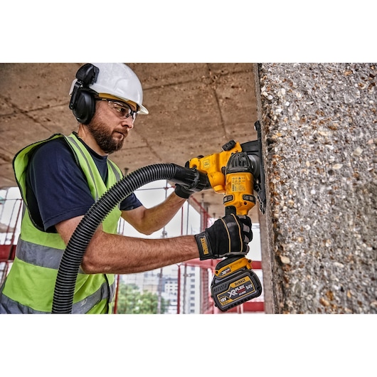 A worker wearing a safety helmet, ear protection, and high-visibility vest uses a DEWALT DCG200T2 cordless power tool for grinding concrete on a construction site. The worker's face is blurred for privacy.