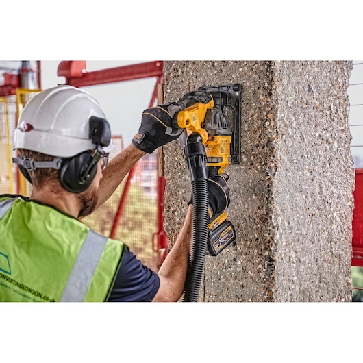 A worker wearing a safety helmet, gloves, and a high-visibility vest uses a DEWALT cordless tool to cut or grind into a concrete pillar. The tool appears to be fitted with a dust extraction hose for safety and cleanliness.