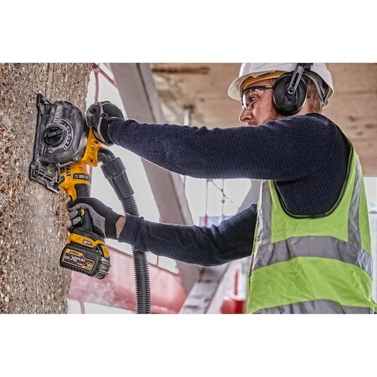 A person wearing safety gear uses a DEWALT cordless wall chaser tool to cut channels into a concrete wall at a construction site.
