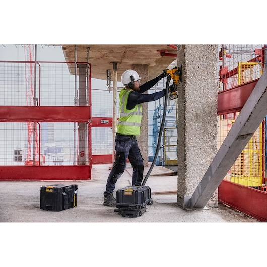 Worker uses DEWALT tool on concrete pillar at construction site, with DEWALT cases and safety gear visible.