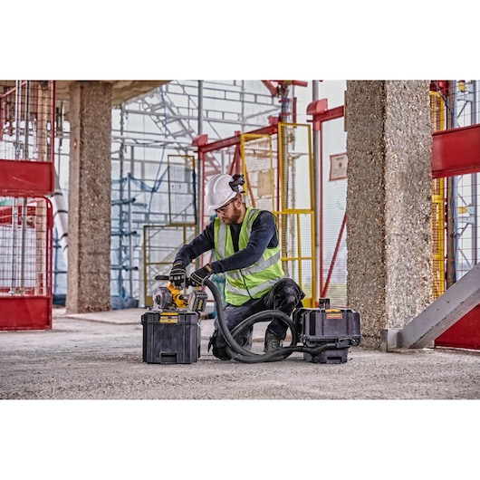 A construction worker wearing a white helmet, gloves, and a reflective safety vest is using a DEWALT power tool connected to dust extraction equipment on a construction site. The worker is kneeling on the ground surrounded by concrete columns and scaffolding.