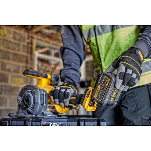 Worker uses DEWALT cordless planer with battery in a construction setting, wearing gloves and safety vest.