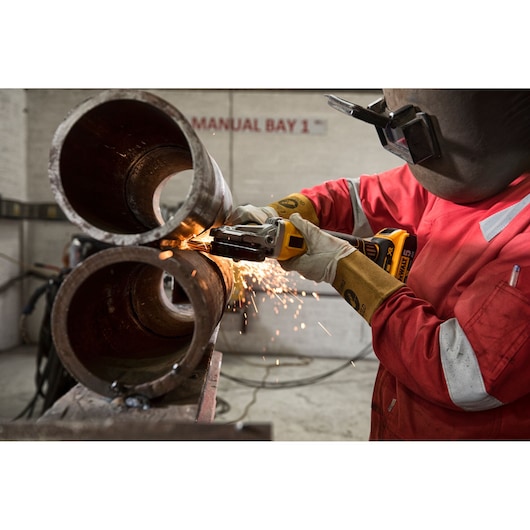 Worker using a DEWALT cordless grinder with sparks on metal pipes in an industrial workshop.