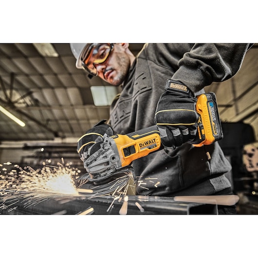DEWALT cordless grinder in use, cutting metal with sparks, worker wearing gloves and safety gear in workshop.