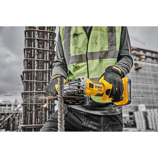 Worker uses a DEWALT cordless grinder to cut rebar on a construction site, wearing gloves and a safety vest.