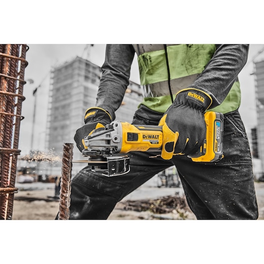 Person uses a DEWALT cordless grinder to cut metal rebar at a construction site, wearing gloves and a safety vest.