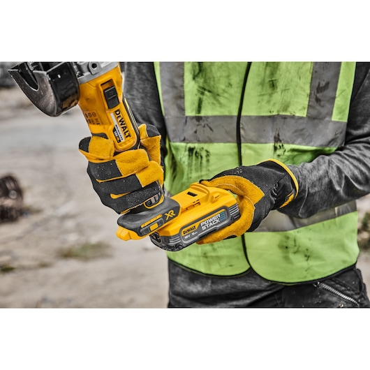 Worker holds DEWALT DCG405H2T grinder with yellow gloves and a green safety vest at a construction site.