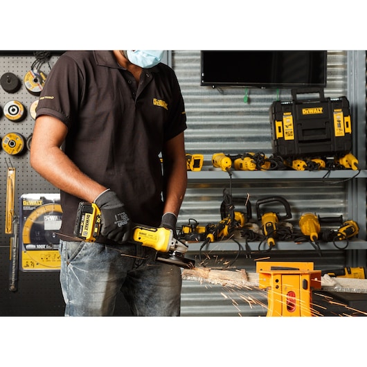 Person uses a DEWALT cordless grinder on metal, wearing gloves and mask. Tools and toolboxes are seen in background.
