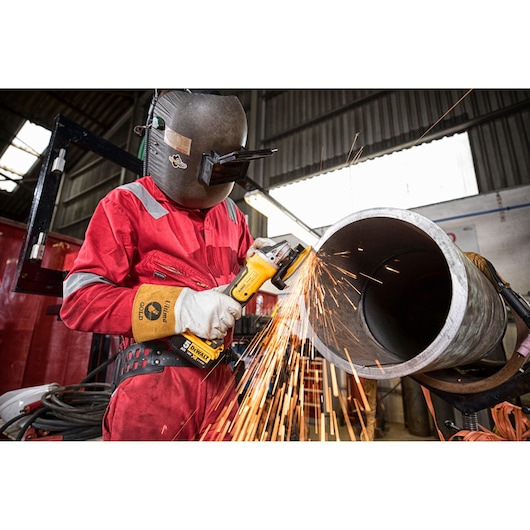 A worker wearing protective gear uses a DEWALT DCG405 cordless angle grinder to cut or grind a metal pipe, with visible sparks flying in an industrial workshop.