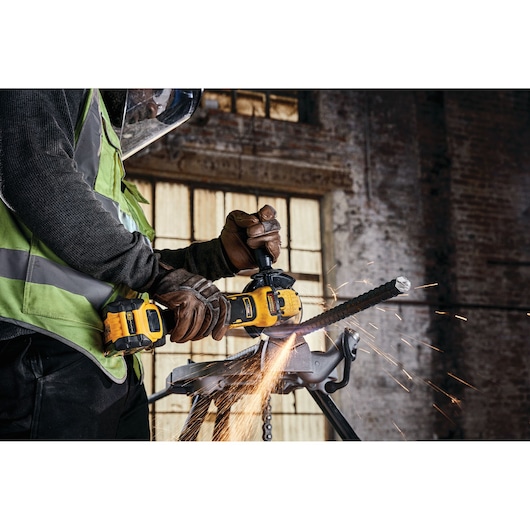Worker uses DEWALT cordless grinder, cutting metal rod with sparks flying in industrial setting.