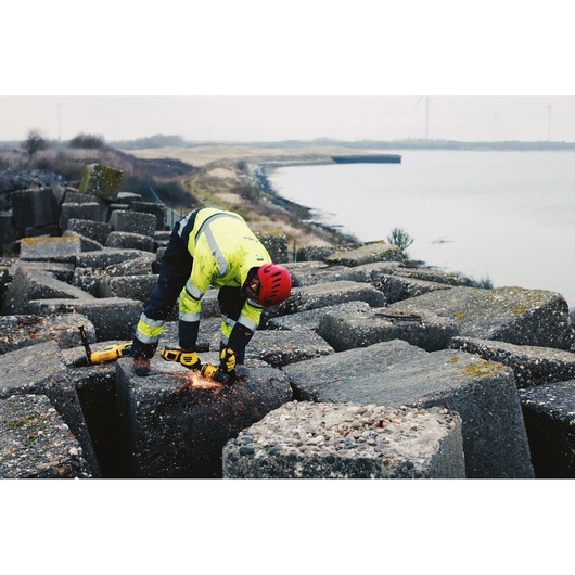 Worker uses DEWALT angle grinder on large concrete blocks by the waterfront, wearing safety gear and helmet.