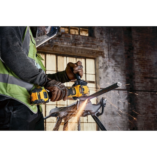 Person uses a DEWALT cordless grinder to cut metal rod, creating sparks in an industrial workshop.