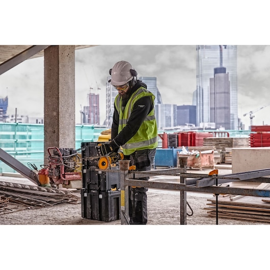 Worker uses DEWALT power tool DCG418T2 on a construction site with city buildings in the background.
