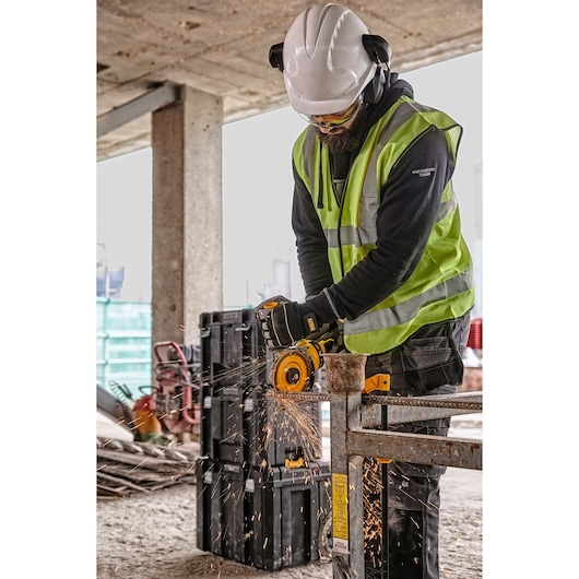 Worker uses DEWALT grinder to cut metal, wearing safety gear on a construction site. Sparks fly from the grinding.
