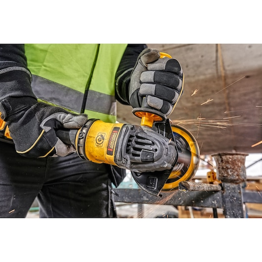 Worker uses yellow DEWALT angle grinder to cut metal, wearing gloves and safety vest. Sparks visible during operation.