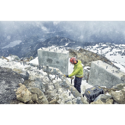 Worker uses DEWALT tool outdoors in snowy mountain area near concrete blocks, with DEWALT case on the ground.