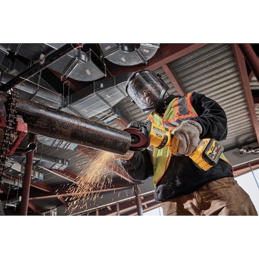 Worker uses DEWALT cordless grinder on metal pipe, wearing safety gear and generating sparks in industrial setting.