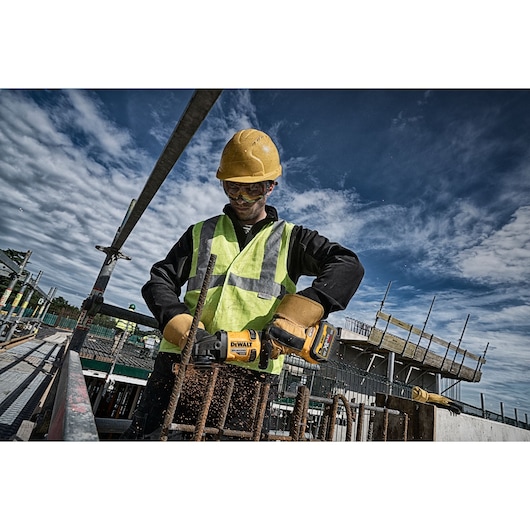 Worker uses DEWALT DCG418T2 grinder to cut metal bars at a construction site, wearing safety gear.