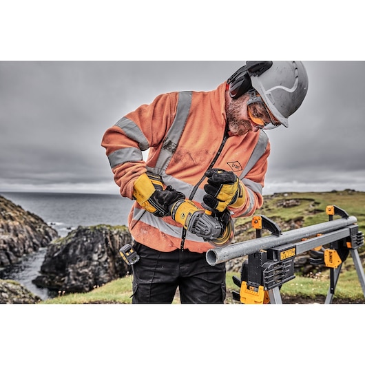 Person using a DEWALT power tool to cut metal pipe outdoors, wearing safety gear and high-visibility clothing.