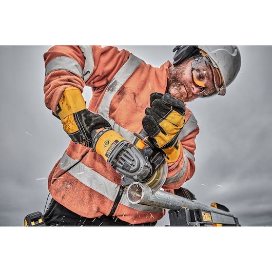Worker uses DEWALT grinder to cut metal pipe, wearing gloves and safety gear outdoors.
