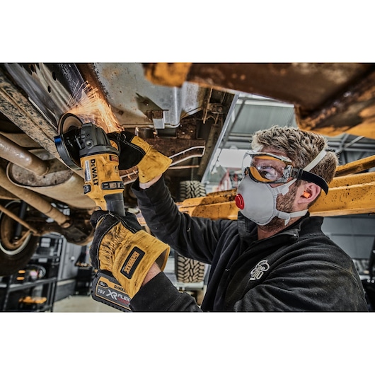 A person wearing safety equipment uses a DEWALT 18V XR FLEXVOLT cordless angle grinder to cut metal underneath a vehicle in a workshop. Sparks are visible where the tool is in contact with the metal.