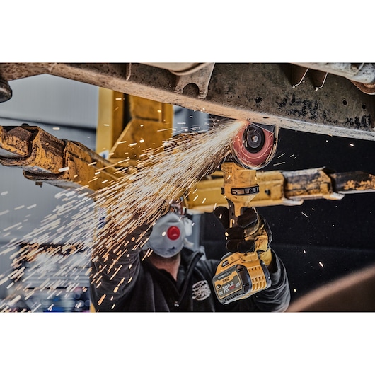 A person wearing safety gloves and a mask is using a DEWALT cordless angle grinder (model DCG418X2) to cut metal, creating sparks in an industrial setting.