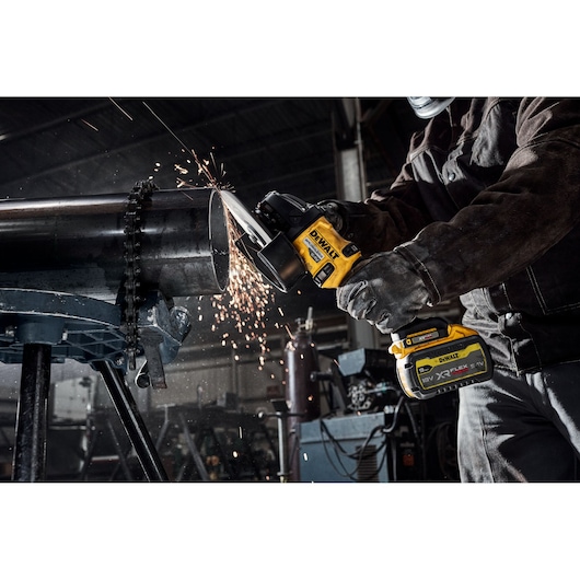 Person uses DEWALT cordless grinder to cut metal pipe, sparks flying in industrial workshop.