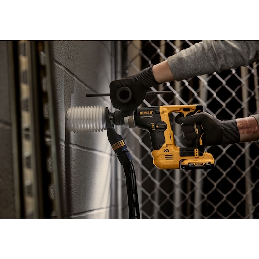 DEWALT cordless rotary hammer drilling into a concrete wall, equipped with a dust extraction attachment.