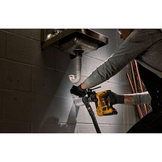 Person uses a DEWALT cordless drill under a sink to work on plumbing against a brick wall.