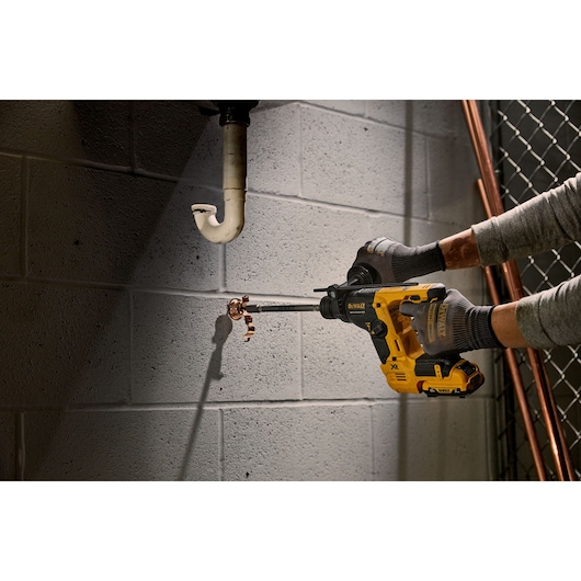 A person uses a DEWALT rotary hammer drill to bore into a concrete wall near plumbing pipes.