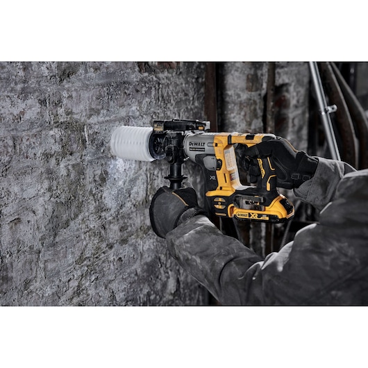 DEWALT cordless hammer drill being used on a concrete wall by a person wearing gloves and a work jacket.