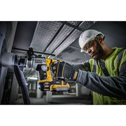 A person wearing a safety helmet and high-visibility vest is using a DEWALT cordless rotary hammer drill to bore into a wall, with dust extraction equipment attached. The drill is labeled with XR and Power Stack branding.