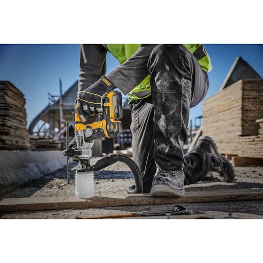 A construction worker is using a DEWALT cordless rotary hammer drill (model DCH172E2T) with dust extraction on a building site, drilling into the ground while kneeling on one knee.