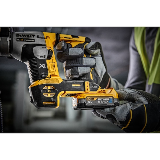 Close-up of a person wearing work gloves holding a DEWALT 18V XR brushless power tool with a Power Stack battery in a workshop or construction environment.