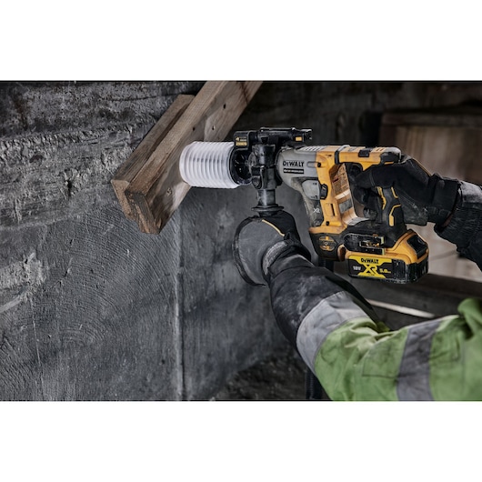 DEWALT cordless rotary hammer drilling into wood and concrete, operated by a gloved worker wearing a green jacket.