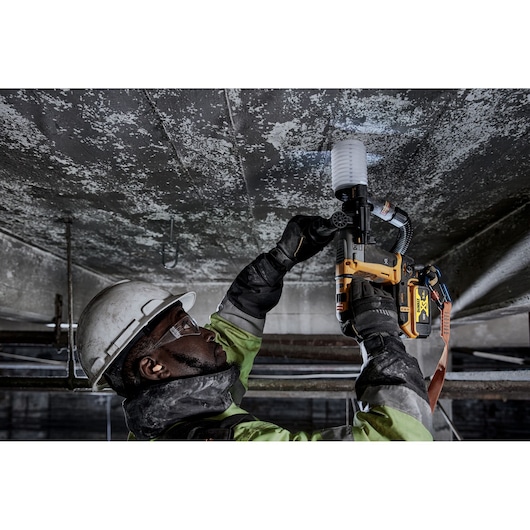 Worker uses DEWALT DCH172P2 rotary hammer drill on a ceiling, wearing safety gear in an industrial setting.