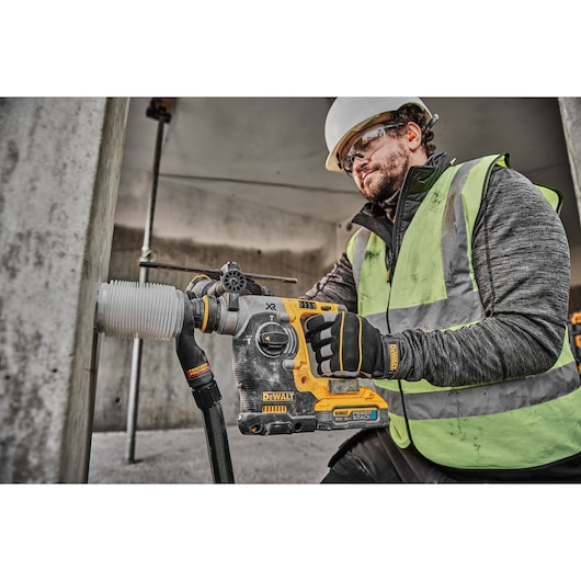 A construction worker wearing a safety helmet and high-visibility vest uses a DEWALT DCH273H2T cordless rotary hammer drill to drill into a concrete column. The worker is also wearing gloves branded with DEWALT.