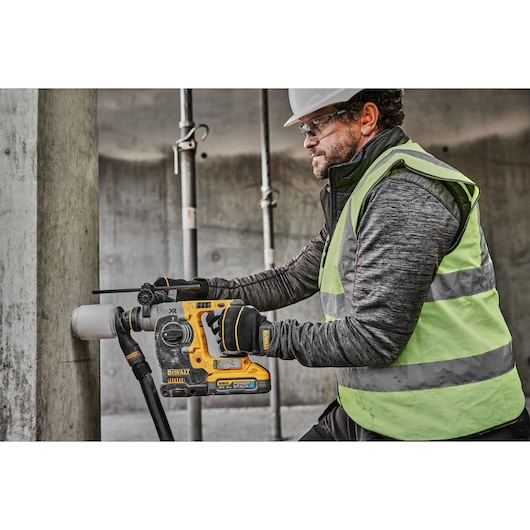 A person wearing a safety vest and gloves is using a DEWALT DCH273H2T cordless rotary hammer drill to drill into a concrete pillar at a construction site. The user's face is blurred for privacy.