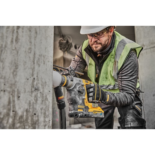 A construction worker using a DEWALT DCH273H2T rotary hammer drill with dust extraction, drilling into concrete. Worker is wearing safety gloves, high-visibility vest, and hard hat. The drill is branded DEWALT and features the PowerStack battery.