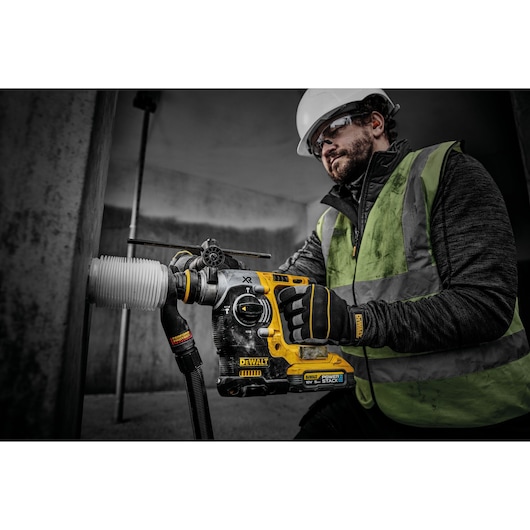 A person wearing a safety vest, gloves, and a hard hat is using a DEWALT DCH273H2T rotary hammer drill to make a hole in a concrete wall. The tool is yellow and black, and connected to a vacuum hose for dust extraction.