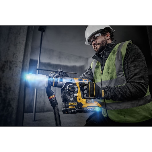 A worker wearing a safety vest, gloves, and a hard hat uses a DEWALT cordless hammer drill to drill into a concrete wall. The drill has a dust extraction attachment and is illuminated with blue lighting, emphasizing the tool's active use and features.