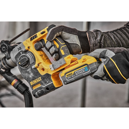 A close-up view of gloved hands holding and installing a DEWALT POWERSTACK 18V battery into a DEWALT cordless rotary hammer drill.