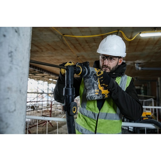 Worker using a DEWALT rotary hammer drill on a construction site, wearing a helmet, gloves, and safety vest.