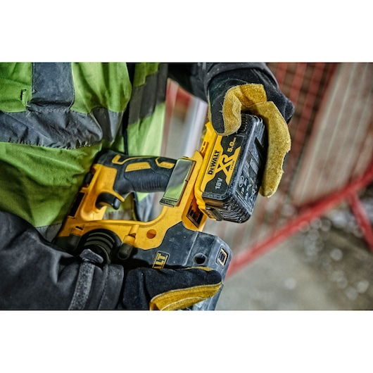 Gloved hand holds a DEWALT DCH274P2T cordless tool with 18V XR battery in a construction setting.